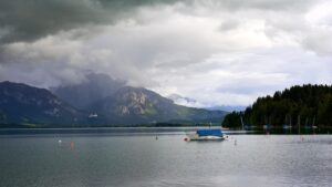 Dieses beeindruckende Foto zeigt den Forggensee an einem bewölkten Tag, eingebettet in die wunderschöne Landschaft der bayerischen Alpen. Der Himmel ist von dichten Wolken bedeckt, die eine dramatische Kulisse für den ruhigen See und die umliegenden Berge schaffen. Ein einzelnes Boot mit einer blauen Plane treibt sanft auf dem Wasser, während rote und weiße Bojen auf der Oberfläche schwimmen. Die üppigen Wälder am Ufer und die schneebedeckten Gipfel im Hintergrund verstärken die natürliche Schönheit dieser Szene. Die friedliche Atmosphäre des Forggensees lädt zum Verweilen und Genießen der Natur ein. Dieses Bild fängt die Essenz der bayerischen Landschaft perfekt ein und bietet einen Moment der Ruhe und Besinnung. Es ist ein idealer Ort für Naturliebhaber und Fotografen, die die wilde Schönheit und die ruhige Gelassenheit der Alpen erleben möchten.